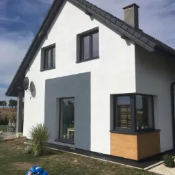 Nowoczesny dom jednorodzinny z białą elewacją, szarą wstawką, ciemnymi oknami i dachem, widok z boku na zadbany trawnik i niebieski worek z odpadami budowlanymi.