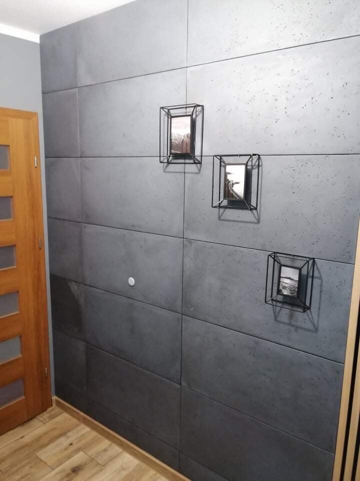 Ściana wykończona panelami imitującymi beton, z trzema metalowymi ramkami z czarno-białymi zdjęciami, drewniane drzwi z przeszkleniami po lewej stronie, parkiet na podłodze.