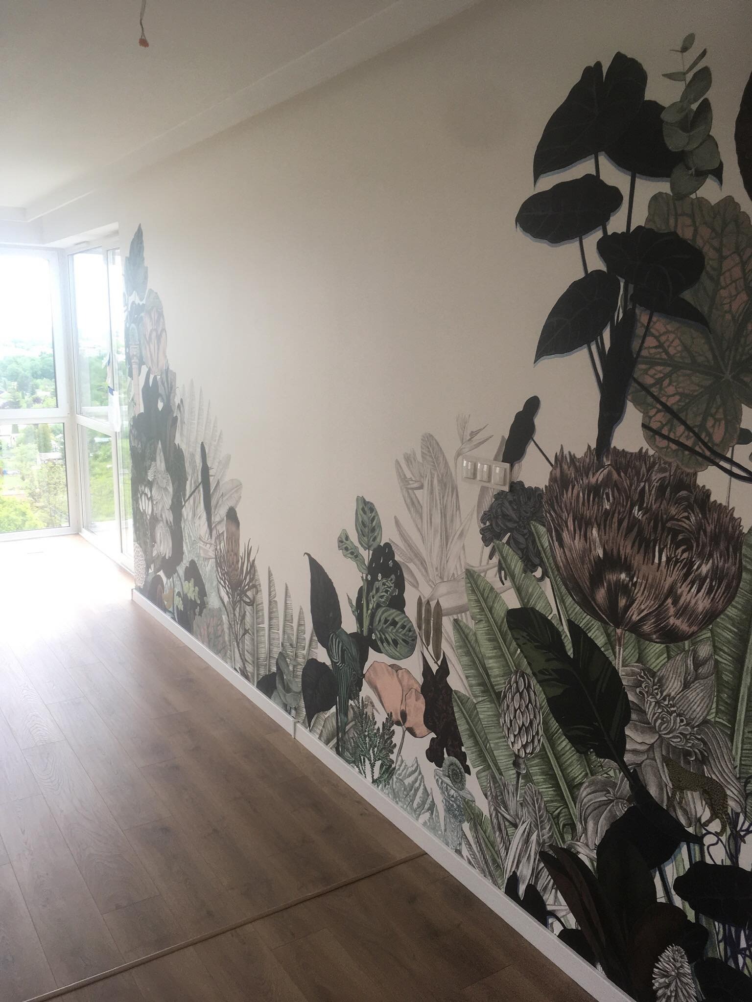 Pokój z parkietem i ścianą ozdobioną fototapetą z motywem tropikalnej roślinności, widok na okno z zielenią za nim.
