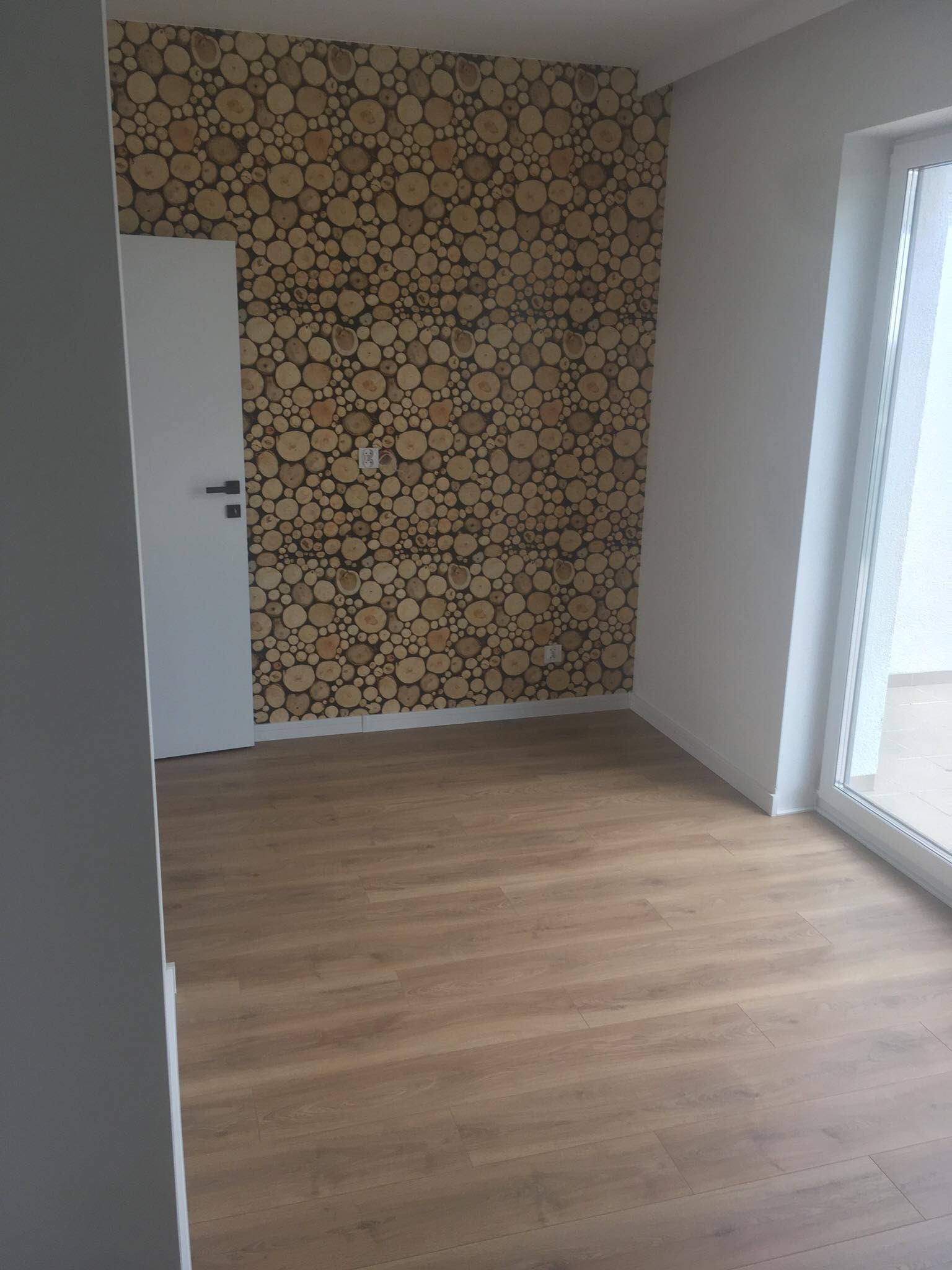 Pokój z drewnianą mozaiką z przekrojów pni na ścianie, jasnymi panelami podłogowymi i białymi drzwiami oraz oknem balkonowym.