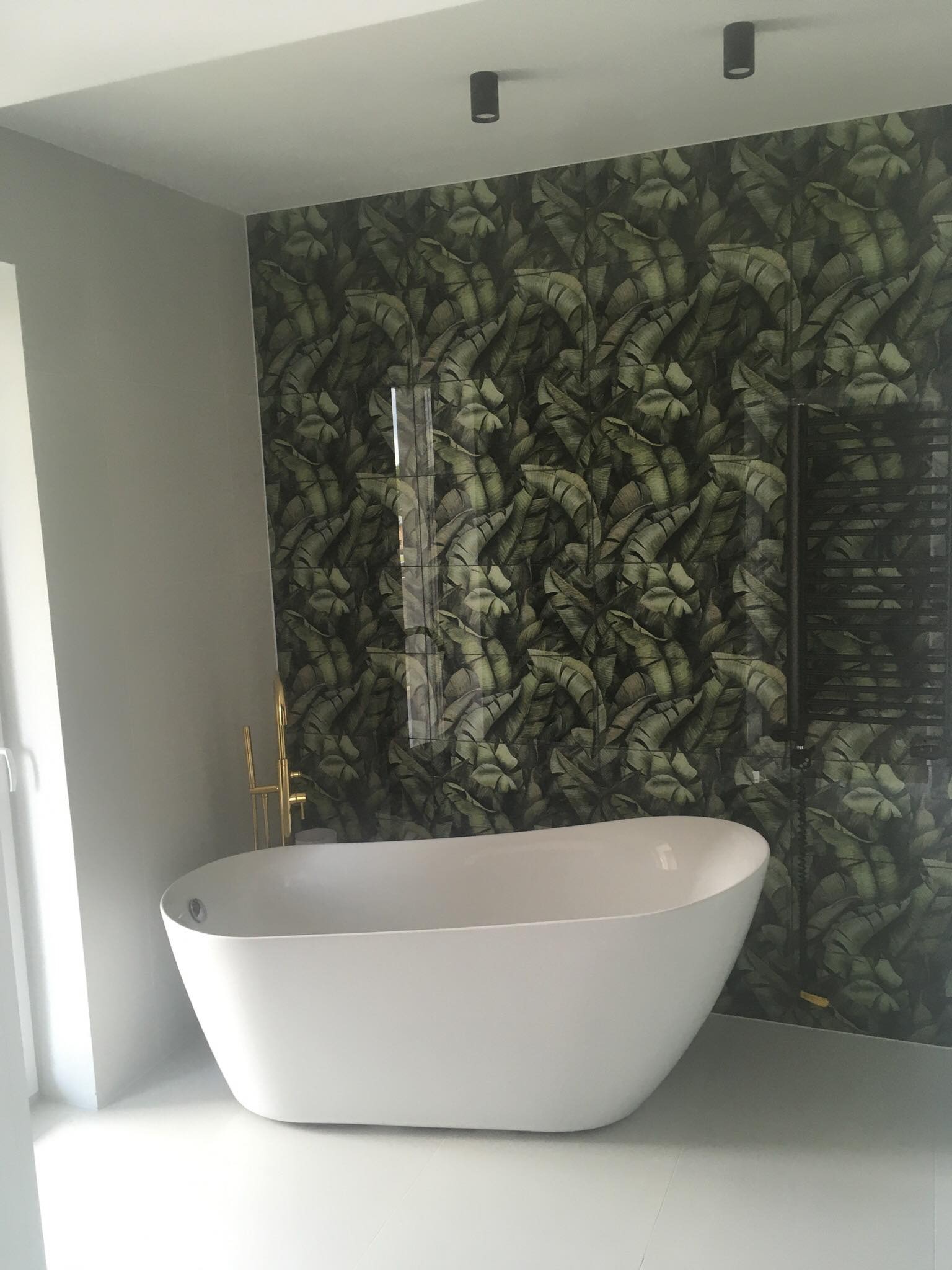 Nowoczesna łazienka z wolnostojącą białą wanną, bateria wannowa w kolorze złotym, ściana wyłożona płytkami z motywem liści bananowca, czarny grzejnik drabinkowy i minimalistyczne oświetlenie sufitowe.
