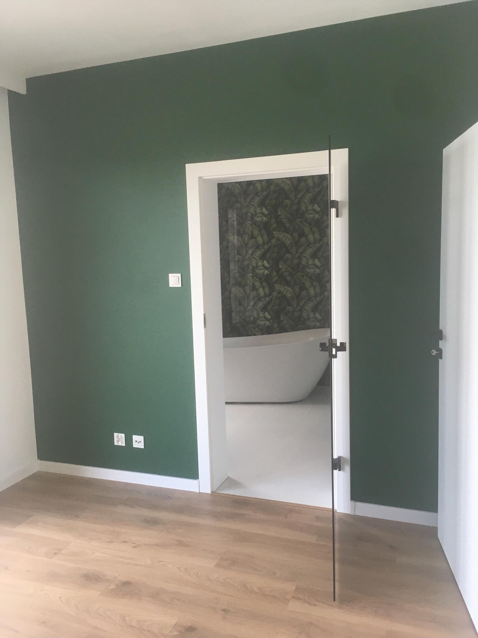 Pokój z zieloną ścianą, drewnianą podłogą i widokiem na łazienkę z wanną przez szklane drzwi.