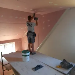 Pracownik w koszulce z logo firmy PNB Dekoracja montuje płytę gipsowo-kartonową do sufitu z folią termoizolacyjną, stojąc na drewnianym podeście, widoczna puszka z masą szpachlową i narzędzia.