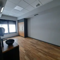 Wnętrze biura z drewnopodobną podłogą, szafą w zabudowie i oknem z żaluzjami, widok na sufit kasetonowy z oświetleniem.