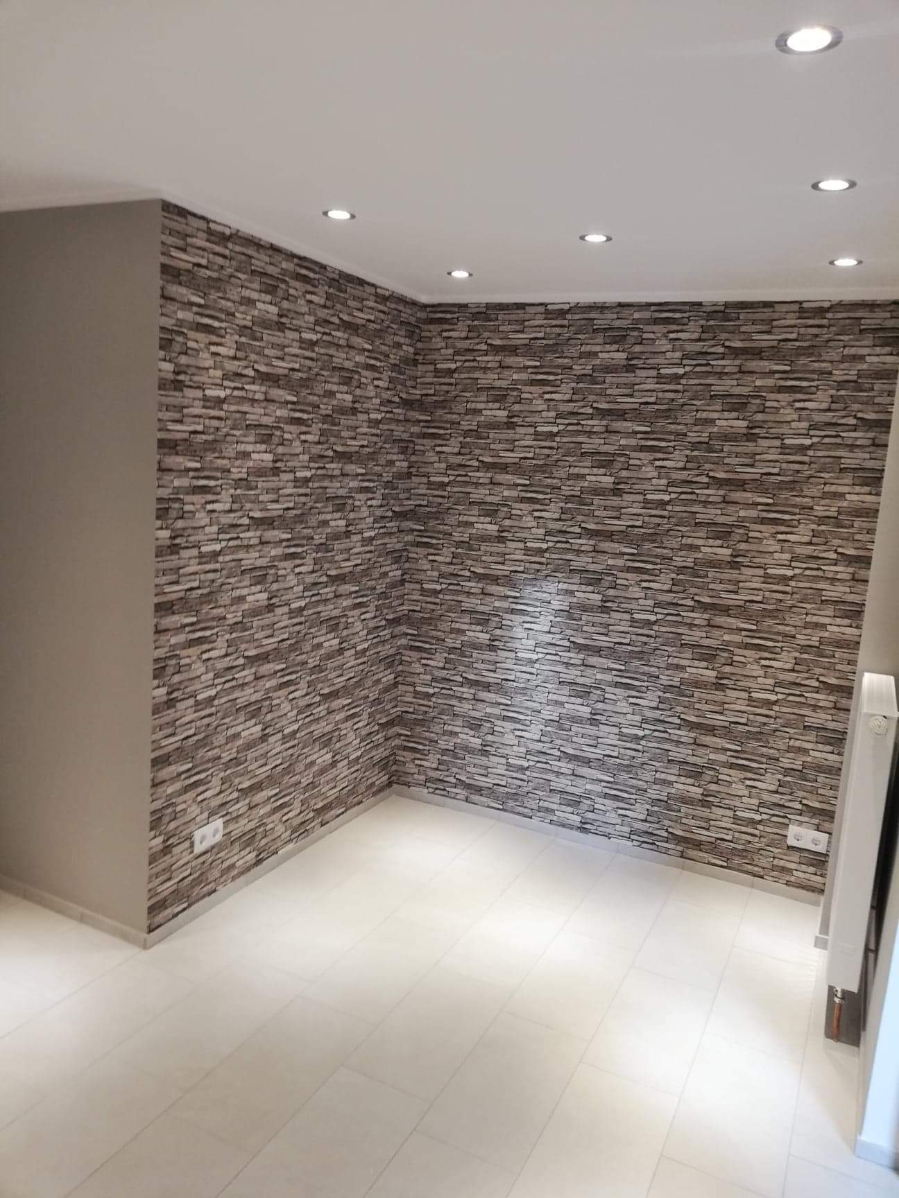 Pusta przestrzeń z wykończoną ścianą imitującą kamień, jasna podłoga z płytek i wbudowane oświetlenie sufitowe.