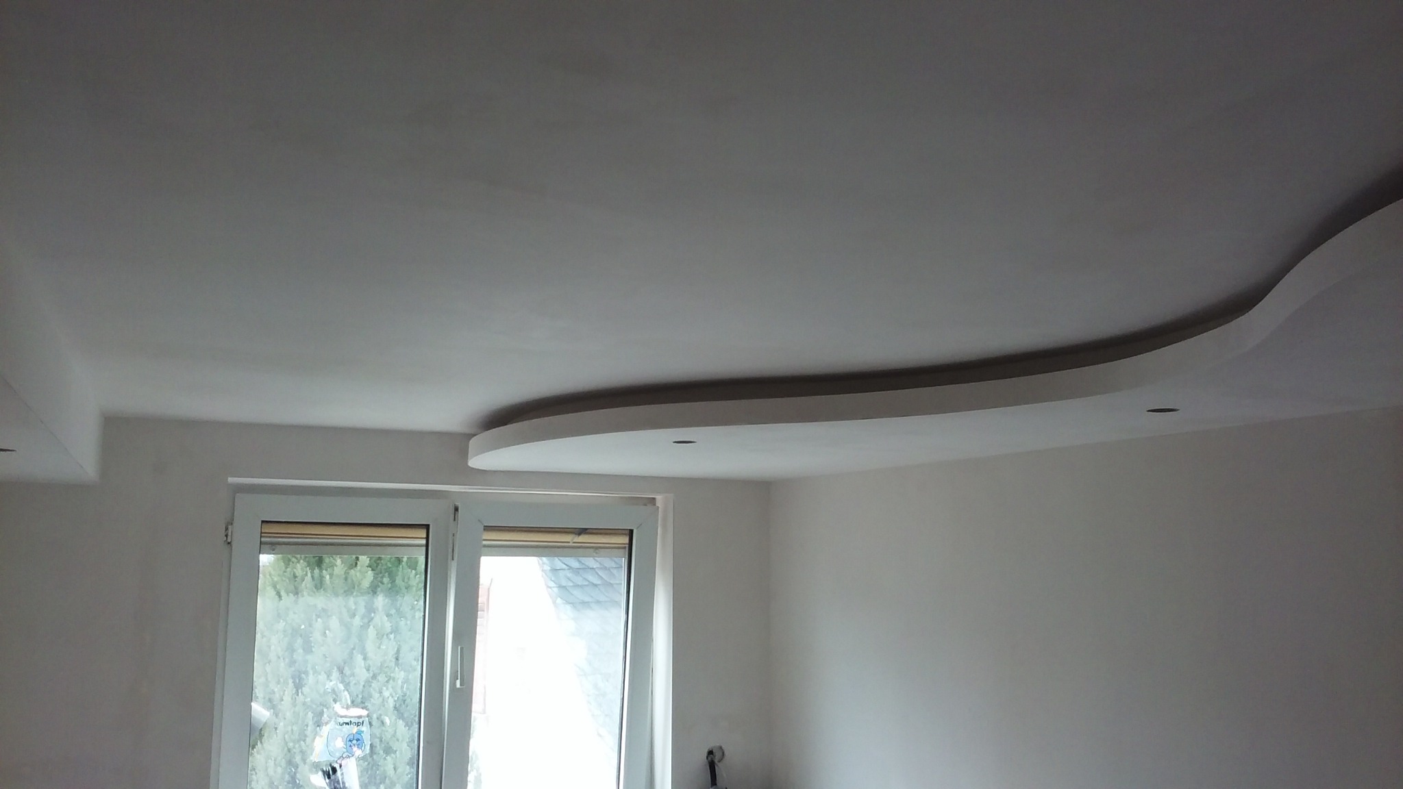 Wnętrze pokoju z gładkimi, pomalowanymi na biało ścianami i sufitem z dekoracyjnym, zaokrąglonym elementem gipsowym z otworem na oświetlenie punktowe, widoczne okno z widokiem na zieleń na zewnątrz.