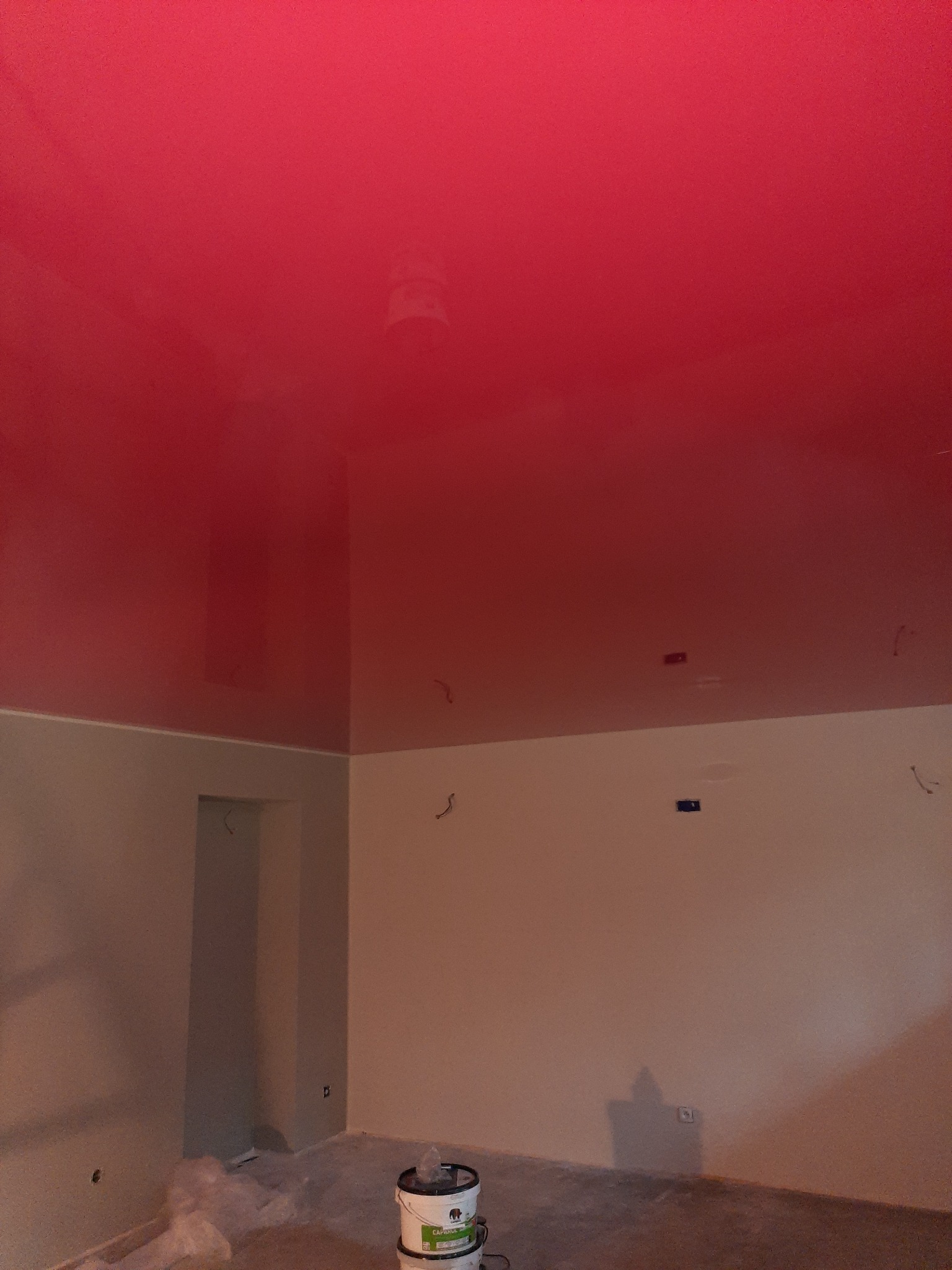 Pomieszczenie w trakcie malowania, z jaskrawoczerwonym, błyszczącym sufitem i ścianami w jasnym odcieniu beżu, widoczny kubeł z farbą Caparol przy ścianie.