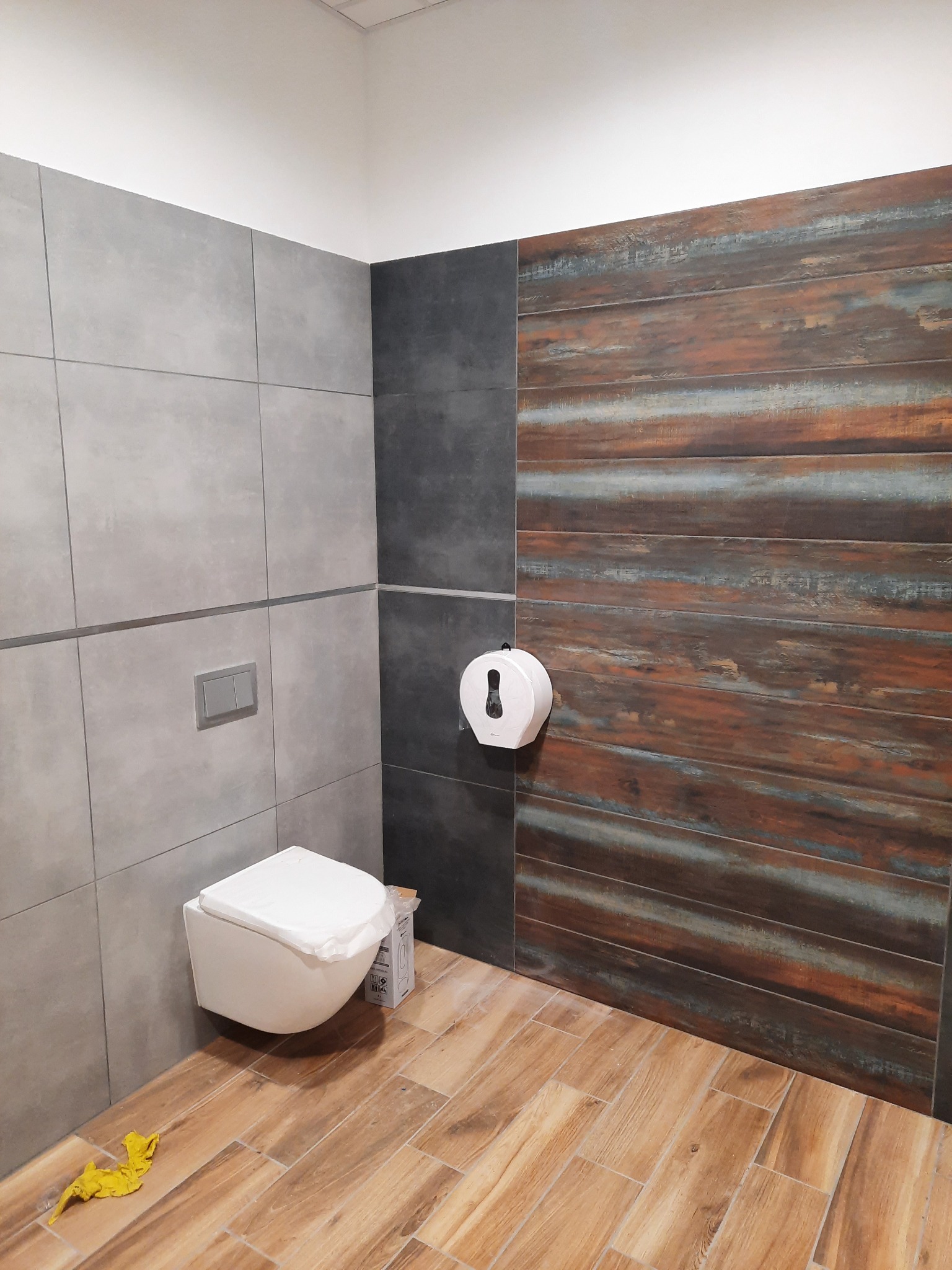Nowoczesna łazienka z wiszącą toaletą, szarą płytką ścienną, akcentem z płytek imitujących drewno i podłogą z drewnopodobnych paneli; widoczny dozownik papieru i żółta ścierka na podłodze.