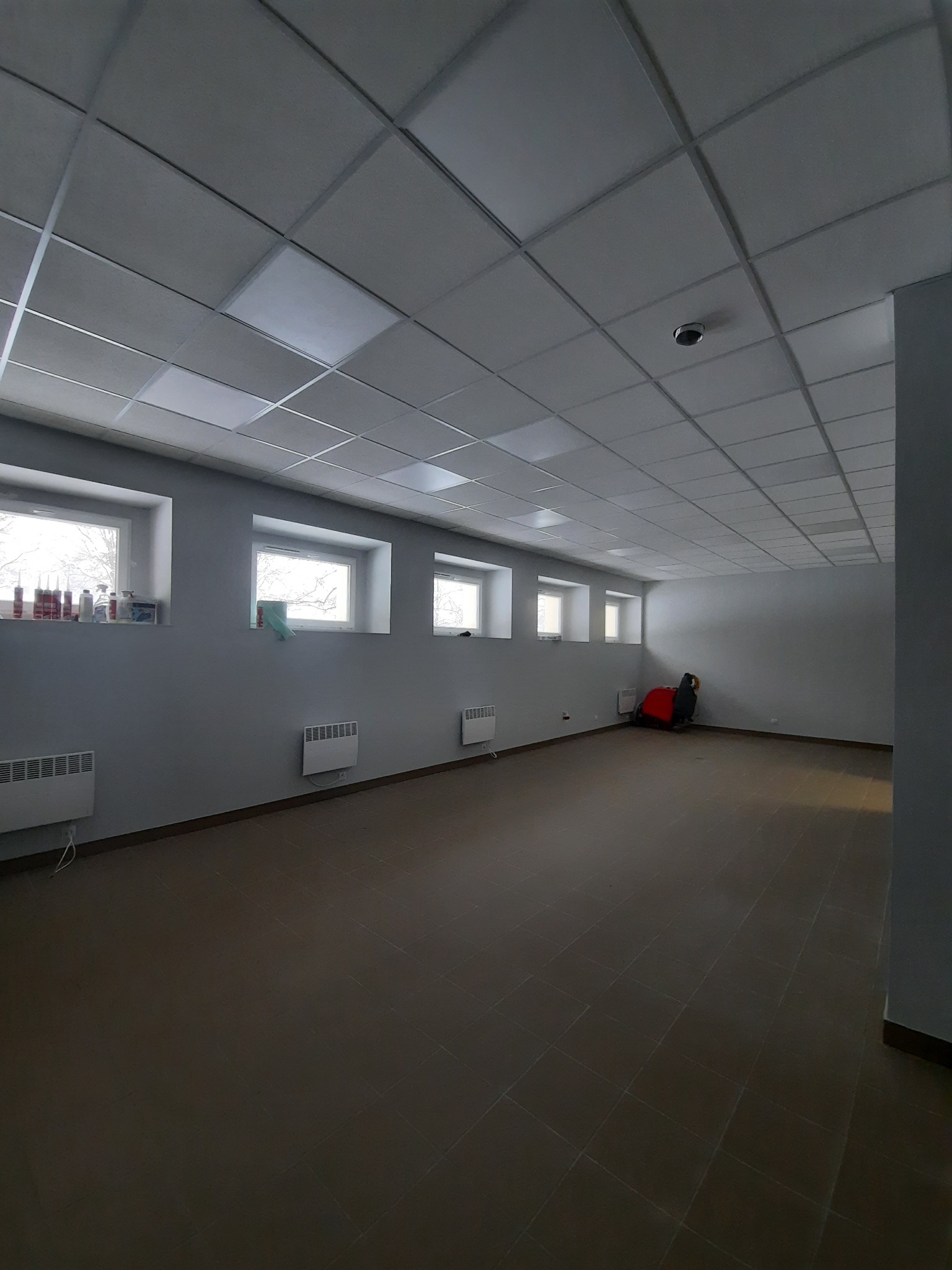 Puste pomieszczenie biurowe z szarą ścianą, brązową podłogą z płytek, oknami z parapetami na których stoją pojemniki, grzejnikami i podwieszanym sufitem z oświetleniem.