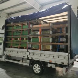 G. S. M. Transport - Biały bus z plandeką załadowany paletami z zielonymi skrzyniami marki Scania, zabezpieczonymi czerwonymi pasami transportowymi, widok z boku na hali.