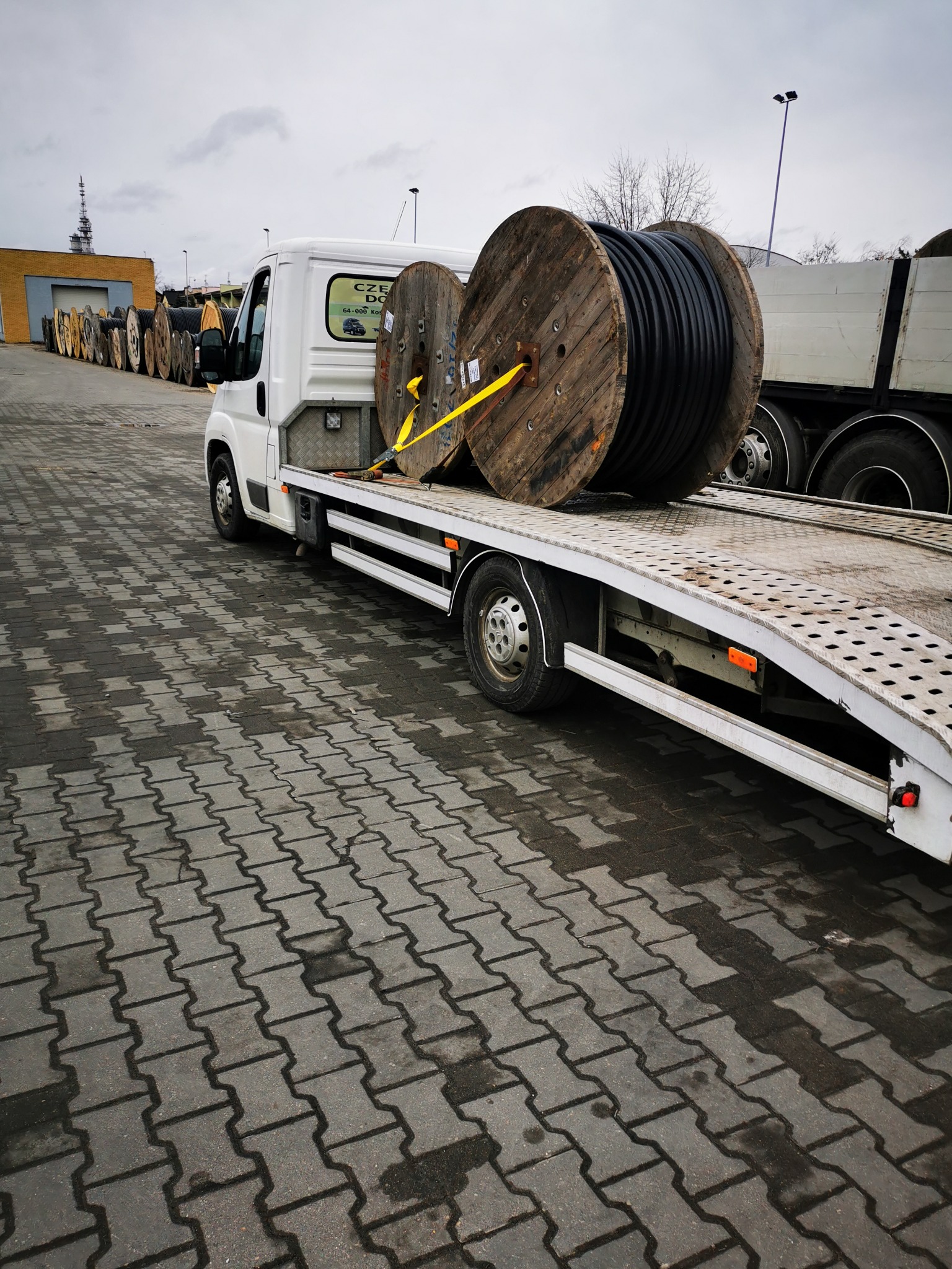 Biała laweta przewozi dwie duże szpule z czarnym kablem, zabezpieczone żółtymi pasami. W tle widoczny rząd szpul i budynek.
