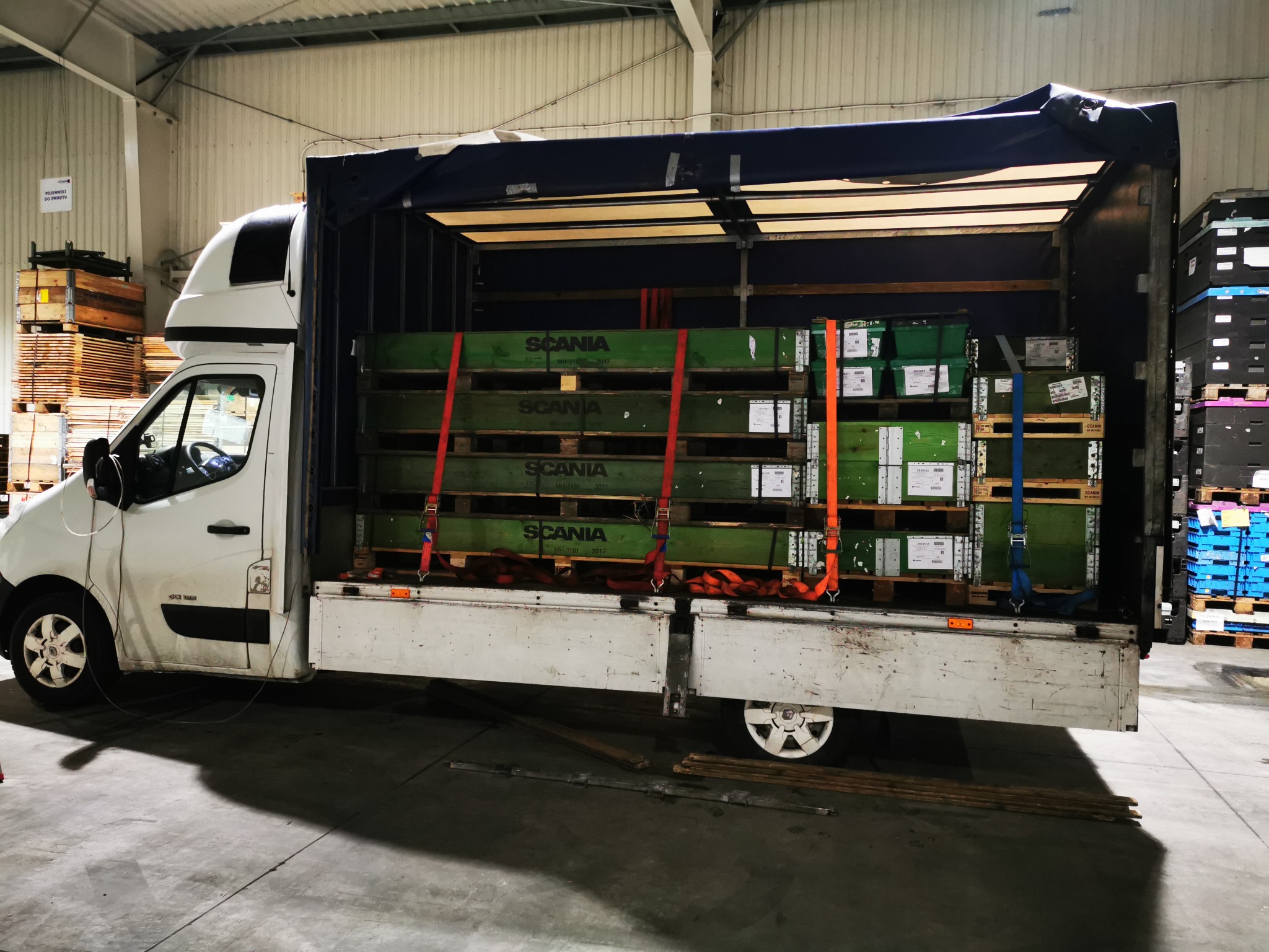 Biały bus z otwartą plandeką załadowany zielonymi skrzyniami z napisem SCANIA, zabezpieczonymi pasami transportowymi w magazynie.