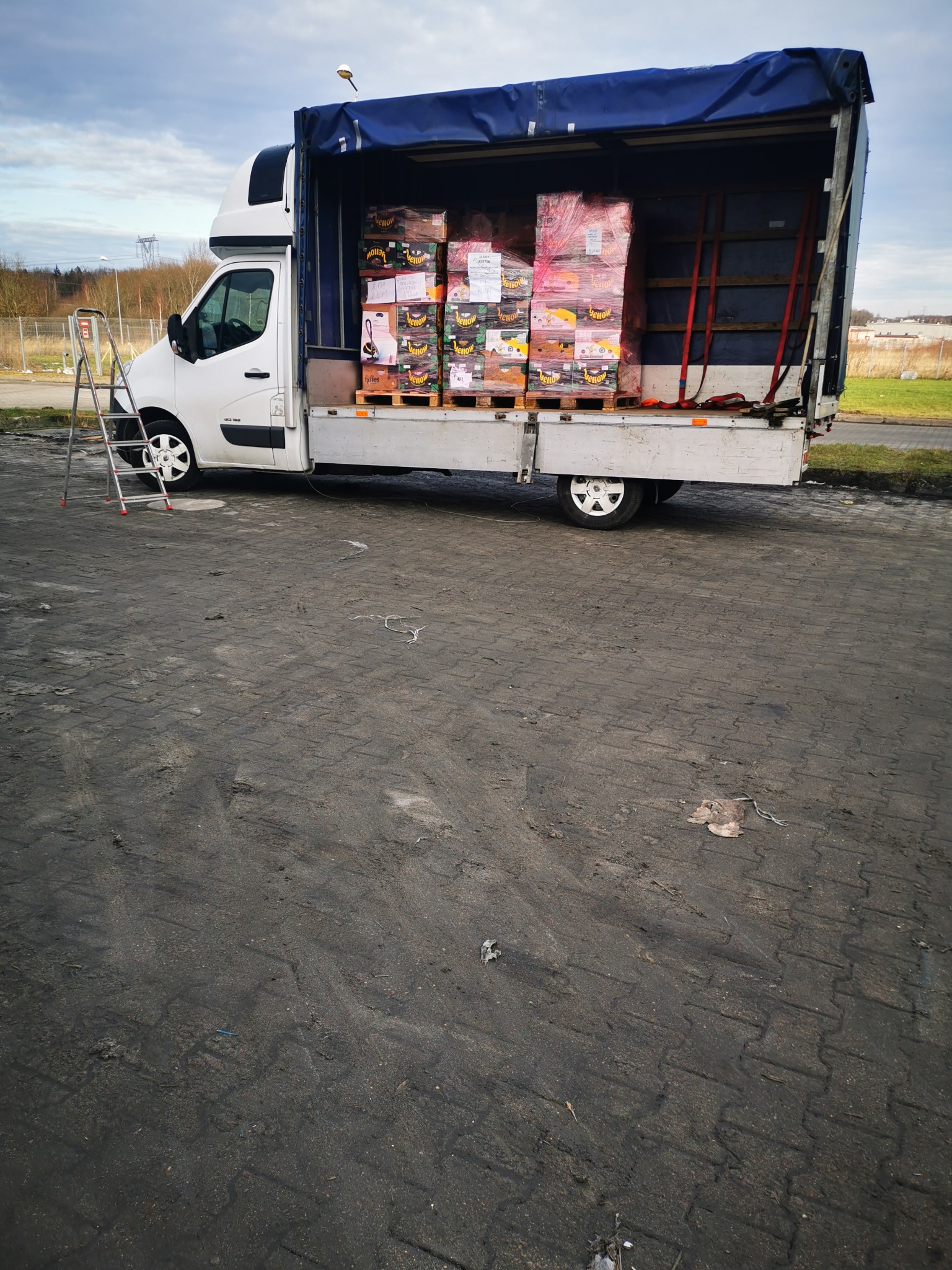 Biały samochód dostawczy z otwartą plandeką, załadowany paletami z kartonami, drabina oparta o bok pojazdu, szaro-bura kostka brukowa na pierwszym planie.