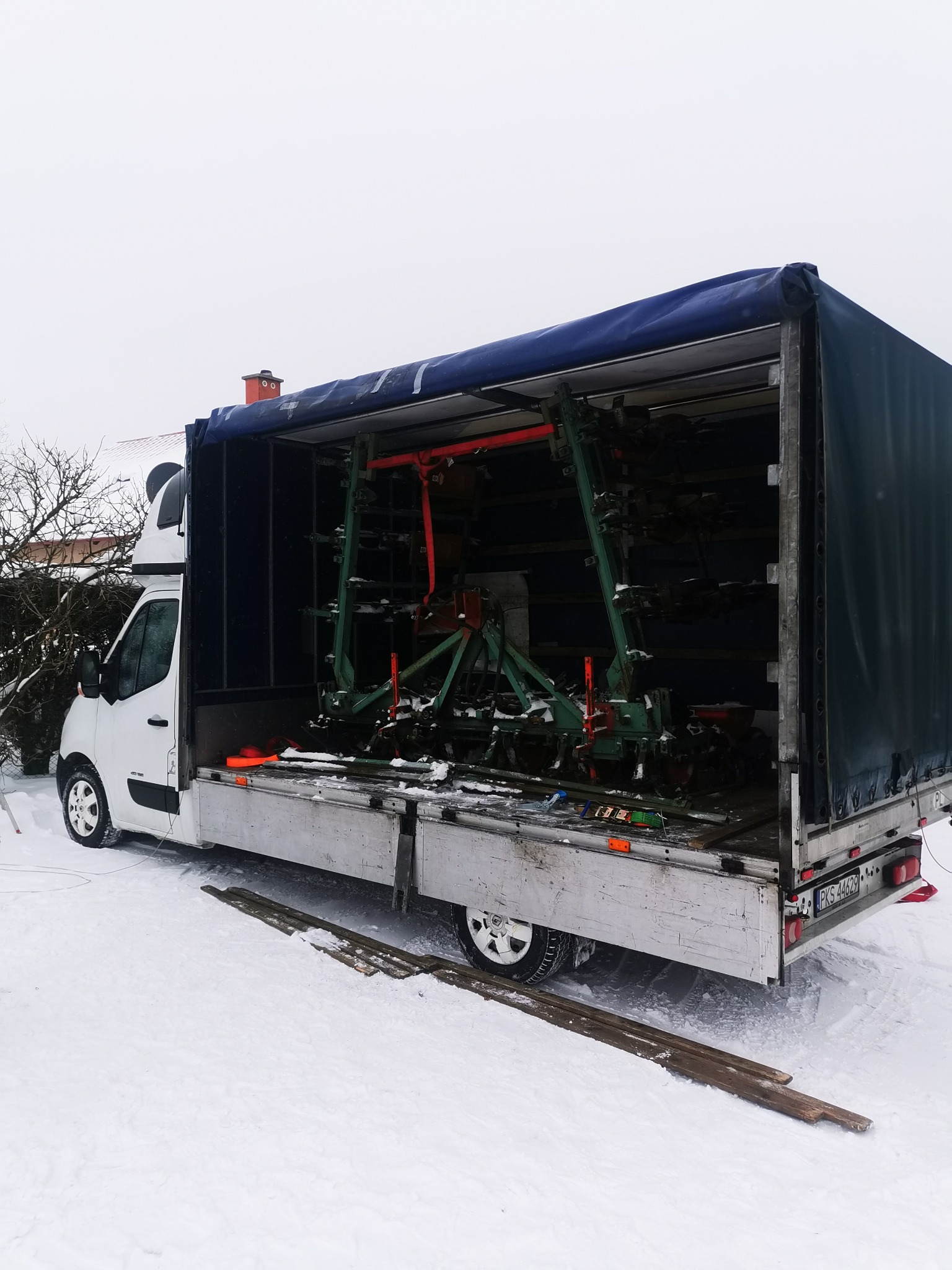 Ciężarówka z otwartą plandeką, załadowana zielonym sprzętem rolniczym, stoi na śniegu w pochmurny dzień.