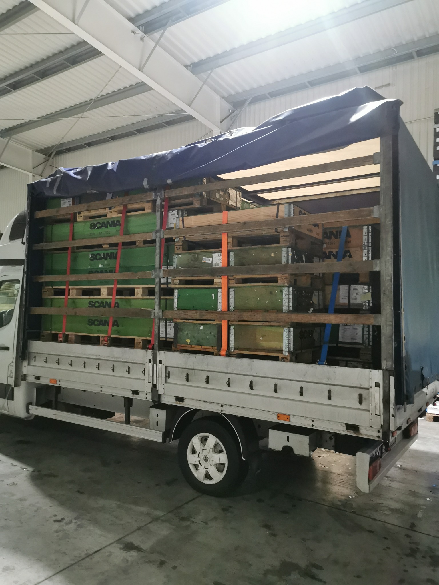 Biały bus z plandeką załadowany paletami z zielonymi skrzyniami marki Scania, zabezpieczonymi czerwonymi pasami transportowymi, widok z boku na hali.