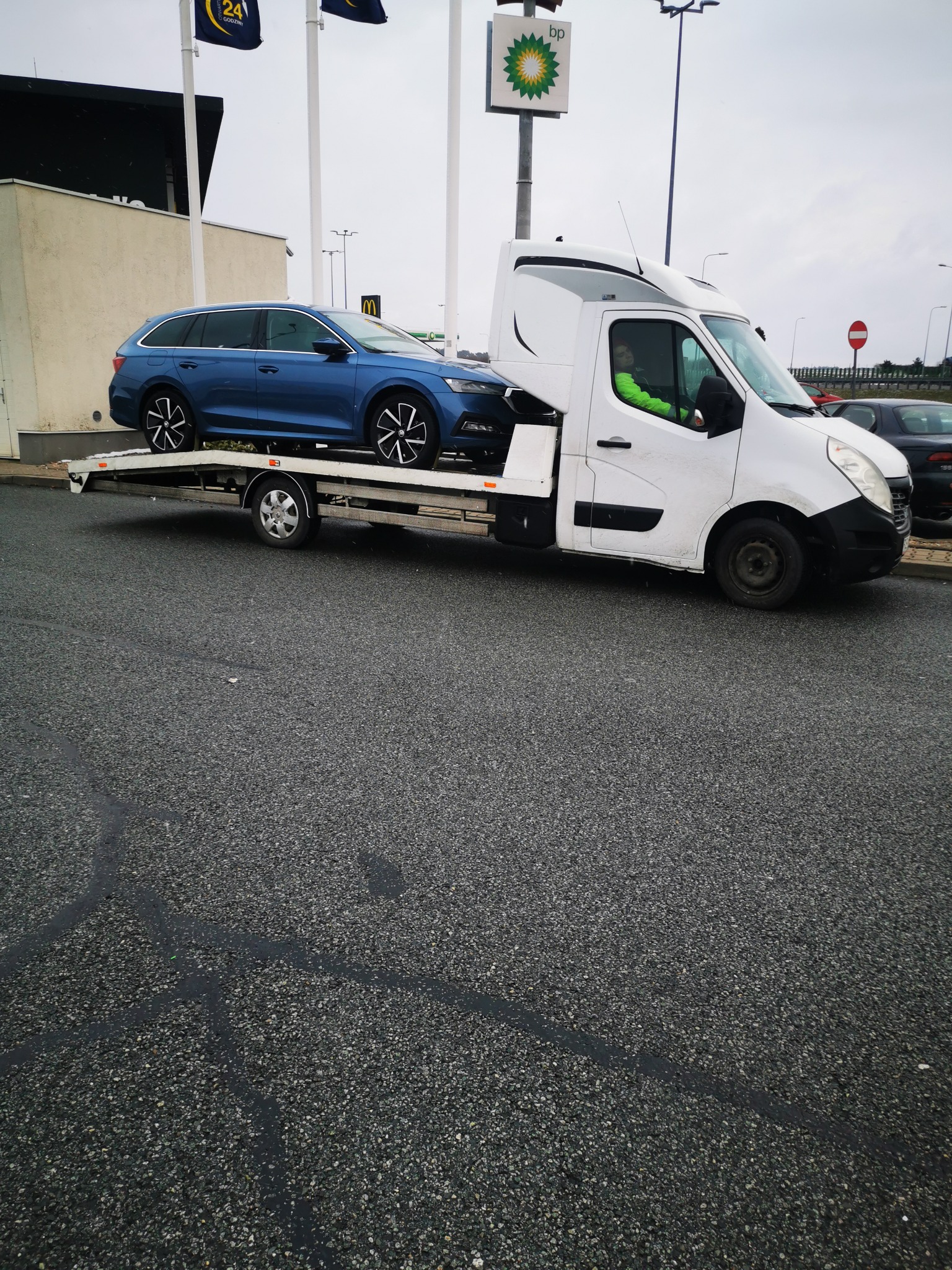 Niebieska Skoda Octavia kombi przewożona na białej lawecie na parkingu stacji benzynowej BP, widoczny kierowca w zielonej kamizelce.
