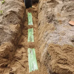 Wykop w piaszczystej glebie z rurą drenarską i zielonymi osłonami filtracyjnymi, widoczny fragment pracownika w tle.