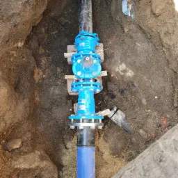 Niebieska armatura z zaworem zamontowana na rurach w wykopie ziemnym, zabezpieczona drewnianymi klockami.