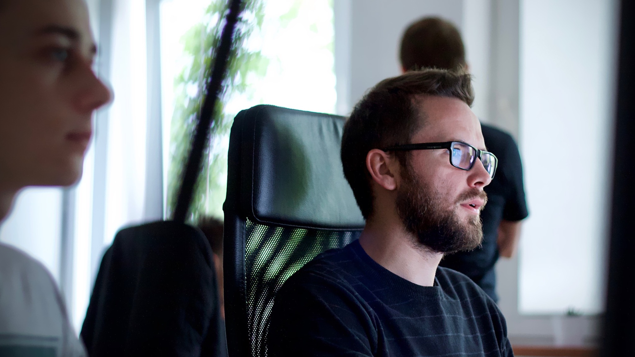 Mężczyzna z brodą i okularami siedzi na krześle biurowym, w tle okno i dwie osoby.
