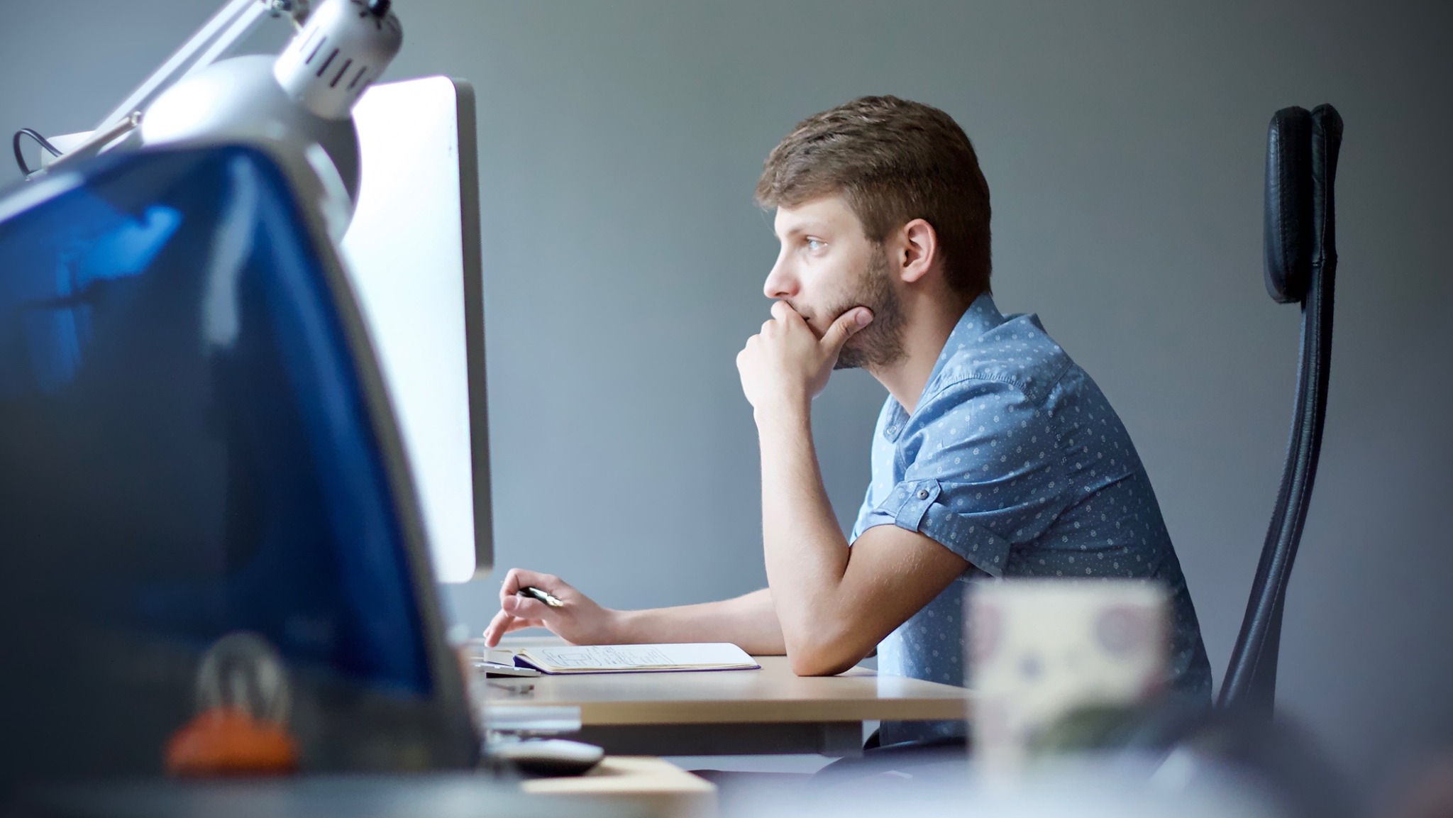 Mężczyzna w błękitnej koszuli zamyślony przed monitorem, notatnik i długopis na biurku, widok z profilu.
