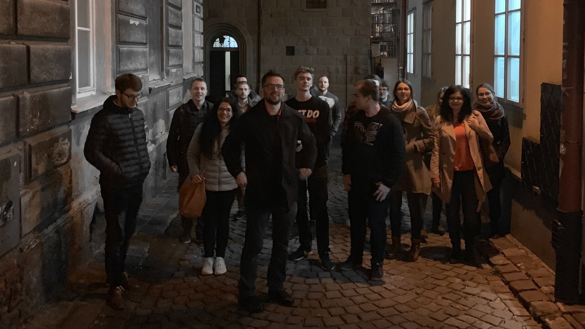 Grupa kilkunastu osób, prawdopodobnie pracowników firmy IT, stoi na brukowanej ulicy w wąskiej uliczce miejskiej, oświetlonej światłem z okien i lamp ulicznych.