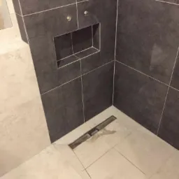 Wykończona kabina prysznicowa z ciemnoszarą glazurą, wnęką ścienną i odpływem liniowym w jasnych płytkach podłogowych.