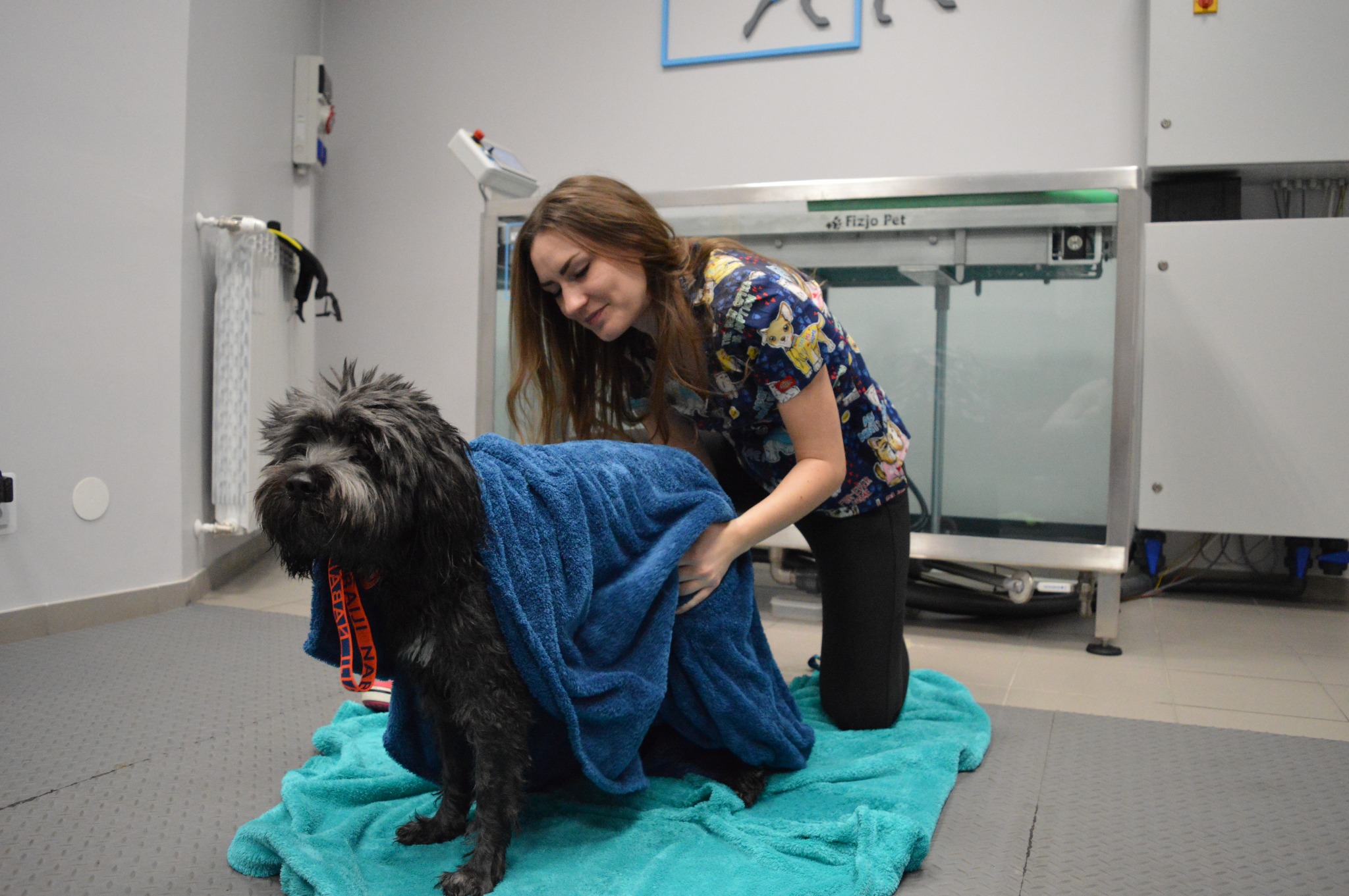 Kobieta owija mokrego, czarnego psa w niebieski ręcznik po hydroterapii, w tle widoczny sprzęt do hydroterapii dla zwierząt.