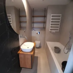 Nowoczesna łazienka z owalną umywalką na drewnianej szafce, czarną ścianą z płytek w jodełkę, białą wanną z wiadrami, grzejnikiem drabinkowym i półkami na ścianie.