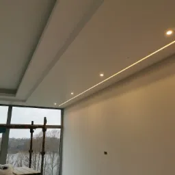 Biały sufit z podwieszanymi elementami i liniowym oświetleniem LED, widok z dołu, w tle okno z rusztowaniem i widokiem na jezioro.