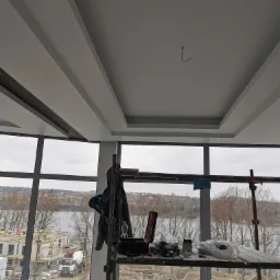 Wnętrze w trakcie wykańczania z widokiem na krajobraz przez duże okna; sufit z dekoracyjnymi elementami, rusztowanie robocze z narzędziami i materiałami w pomieszczeniu.