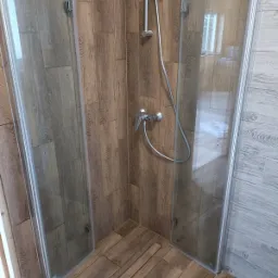 Kabina prysznicowa z drzwiami szklanymi, wykończona płytkami imitującymi drewno na ścianach i podłodze, widoczna bateria i słuchawka prysznicowa.