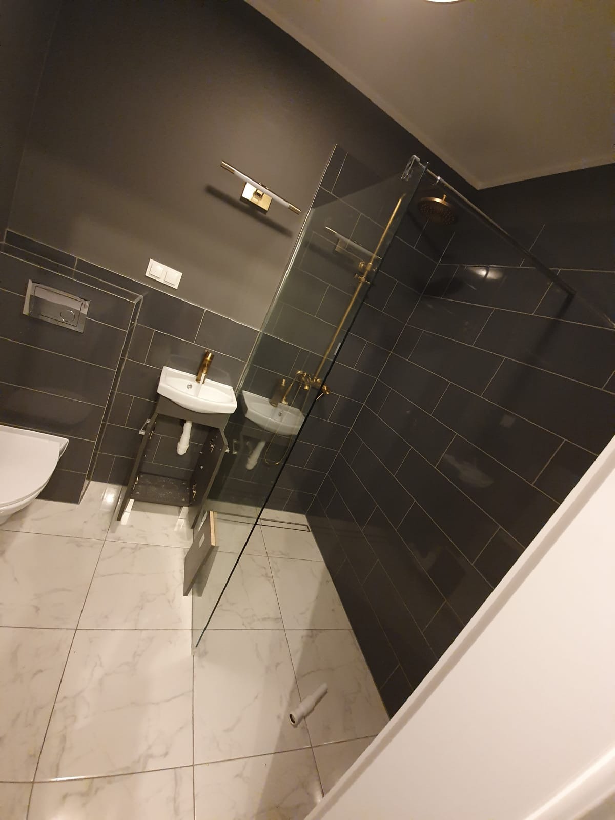 Nowoczesna łazienka z szarymi płytkami, marmurową podłogą i złotą armaturą w kabinie prysznicowej typu walk-in oraz małą umywalką.