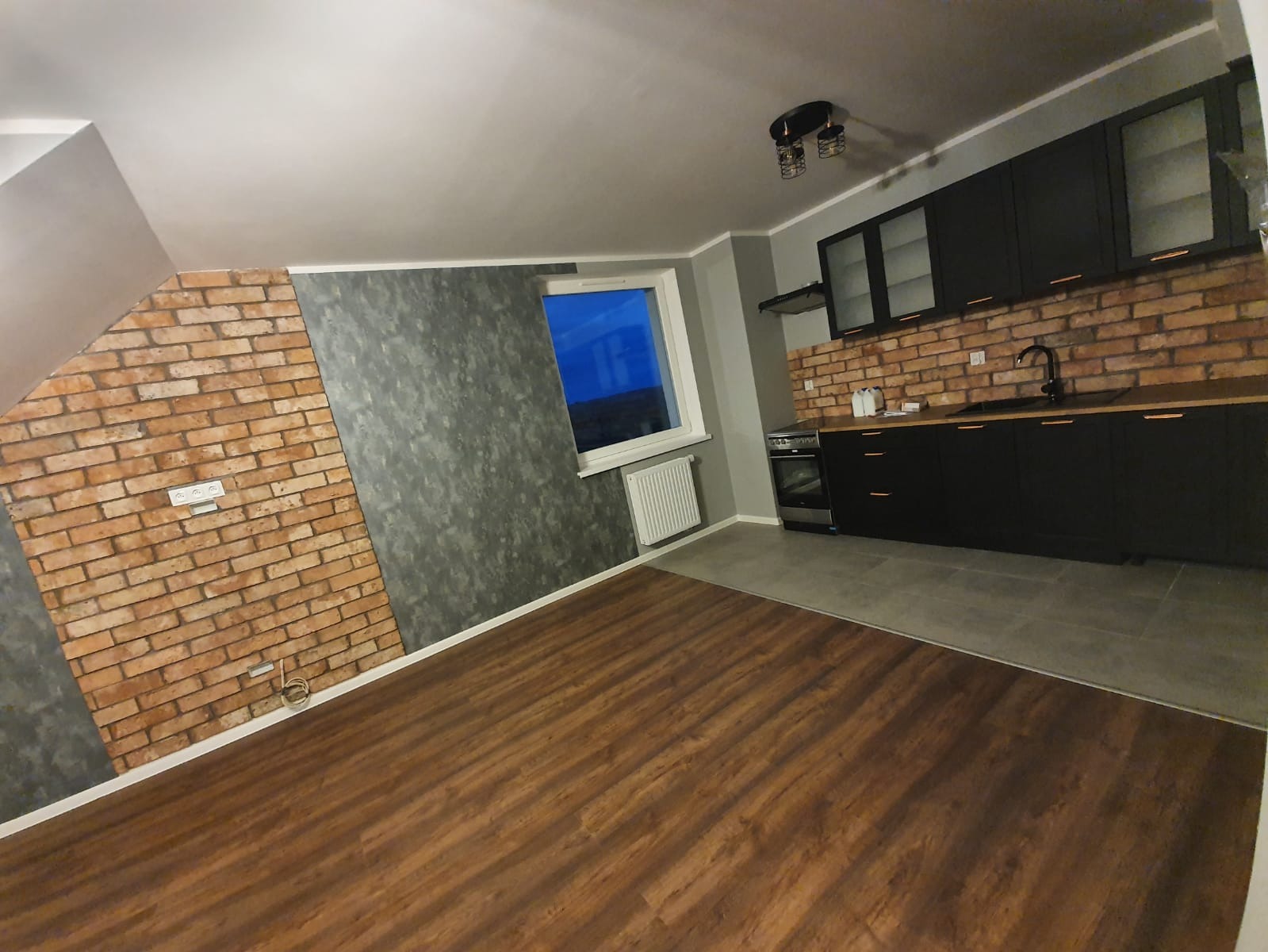Ukończone wnętrze kuchni w odcieniach brązu, szarości i czerni z ceglaną ścianą, ciemnymi meblami kuchennymi i drewnianą podłogą, widok na okno z grzejnikiem.