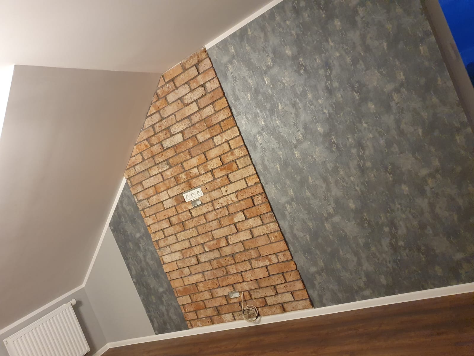 Ściana w pokoju na poddaszu z dekoracyjnym panelem z cegły, obok szara ściana z efektem betonu, widoczne gniazdka elektryczne i fragment grzejnika.