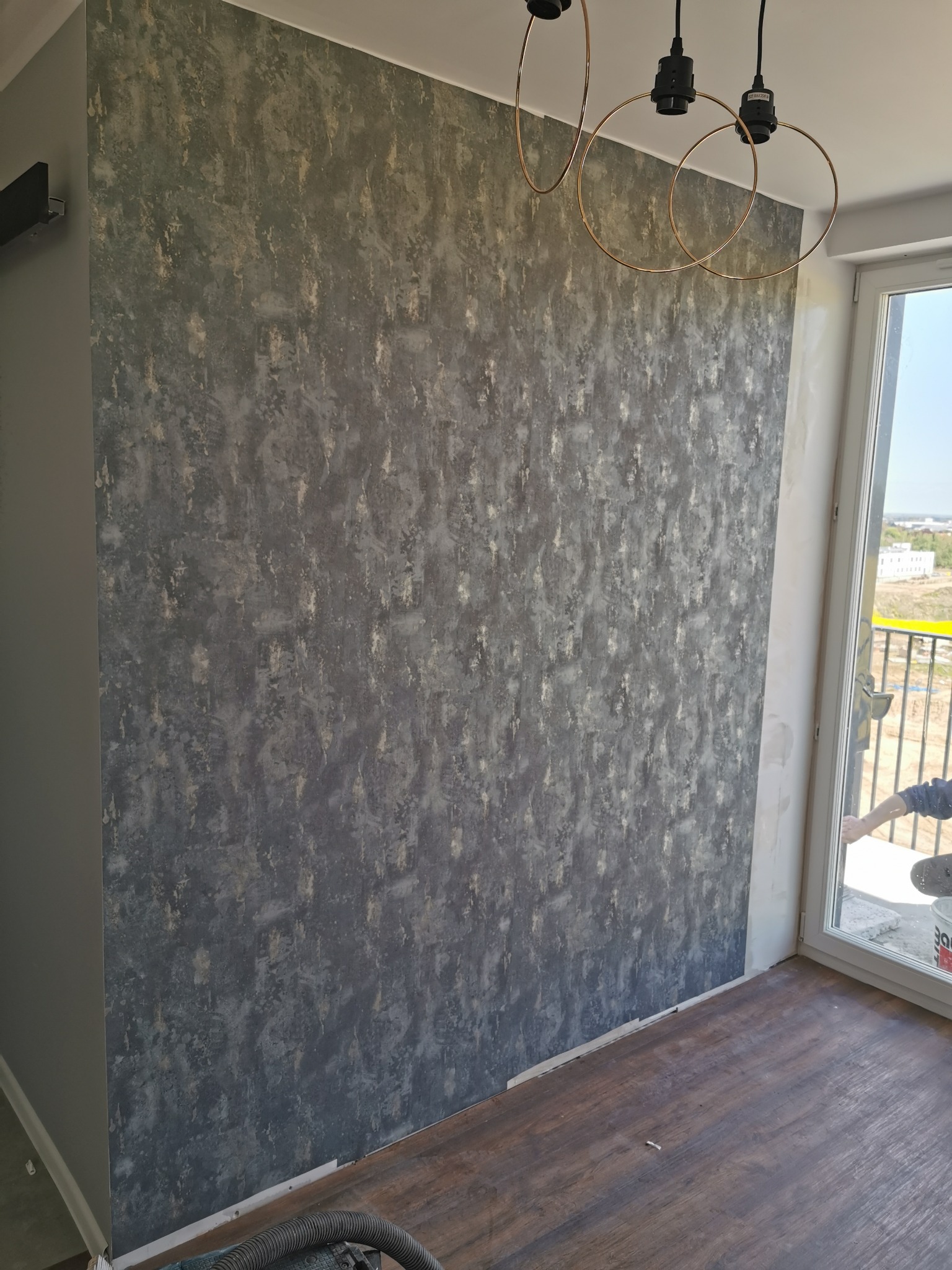 Ściana z dekoracyjną fakturą w odcieniach szarości i złota, oświetlona nowoczesną lampą z okrągłymi kloszami, widoczny fragment okna i podłogi z paneli imitujących drewno, w rogu fragment odkurzacza.