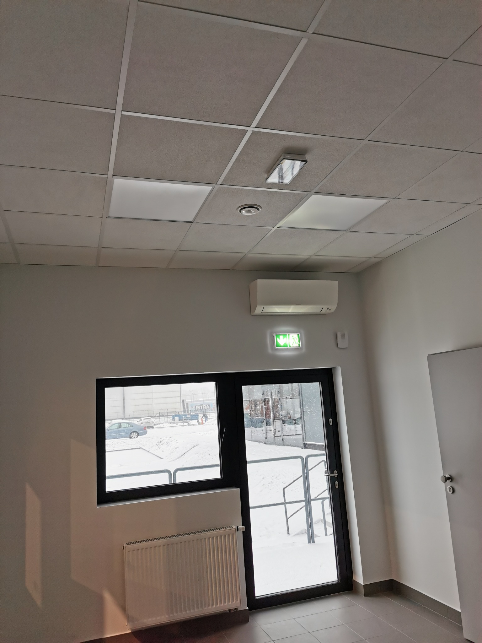 Wnętrze biura po remoncie: białe ściany, sufit kasetonowy z oświetleniem, klimatyzator, okno z widokiem na zimowy krajobraz, grzejnik pod oknem, drzwi ewakuacyjne z zielonym piktogramem.
