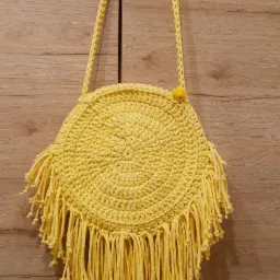 Żółta, okrągła torebka na ramię, wykonana na szydełku z frędzlami, wisząca na drewnianych drzwiach.