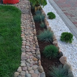 WIOLETTA Wojciechowska - Kaskada ogrodowa z kamienną ścieżką, białym żwirem, ozdobnymi trawami i słonecznikami, zakończona czerwoną biedronką dekoracyjną.