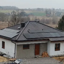 Dom jednorodzinny z zainstalowanymi panelami słonecznymi na dachu z dachówki ceramicznej, widok z góry na tle krajobrazu wiejskiego w pochmurny dzień.