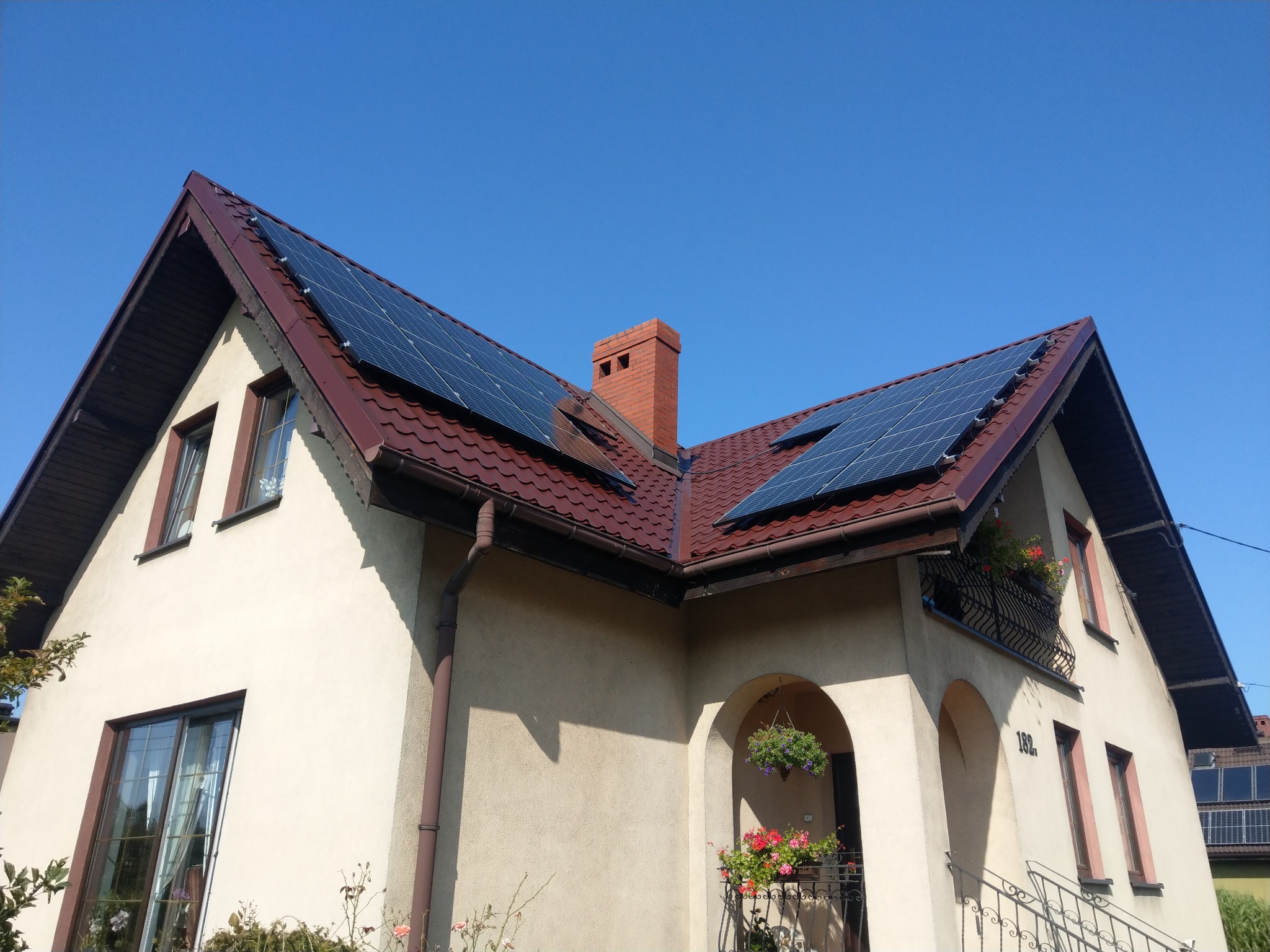 Dom jednorodzinny z panelami słonecznymi na dachu z czerwonej dachówki, widok z dołu w słoneczny dzień, na elewacji kwiaty w donicach.