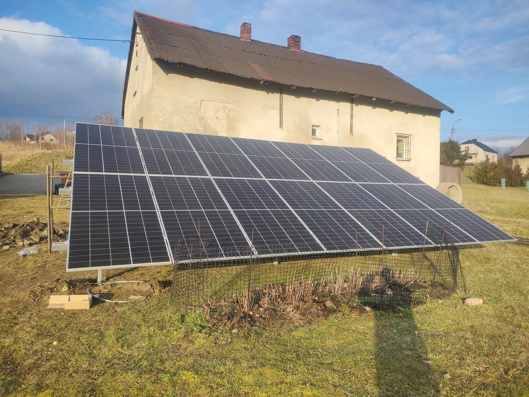 Panele słoneczne na trawniku przed domem z ceglanym dachem, widoczne kable i karton po instalacji.