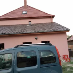 Dwukondygnacyjny budynek z różową elewacją i dwoma rodzajami dachówki, widoczny zza zaparkowanego, niebieskiego samochodu.