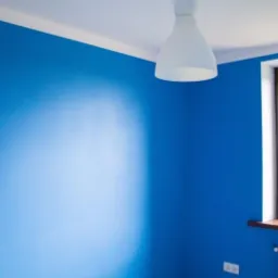 Pokój po remoncie: ściany pomalowane na intensywny niebieski kolor, biały sufit z lampą w kształcie dzwonu, okno z brązową ramą i grzejnik pod nim.