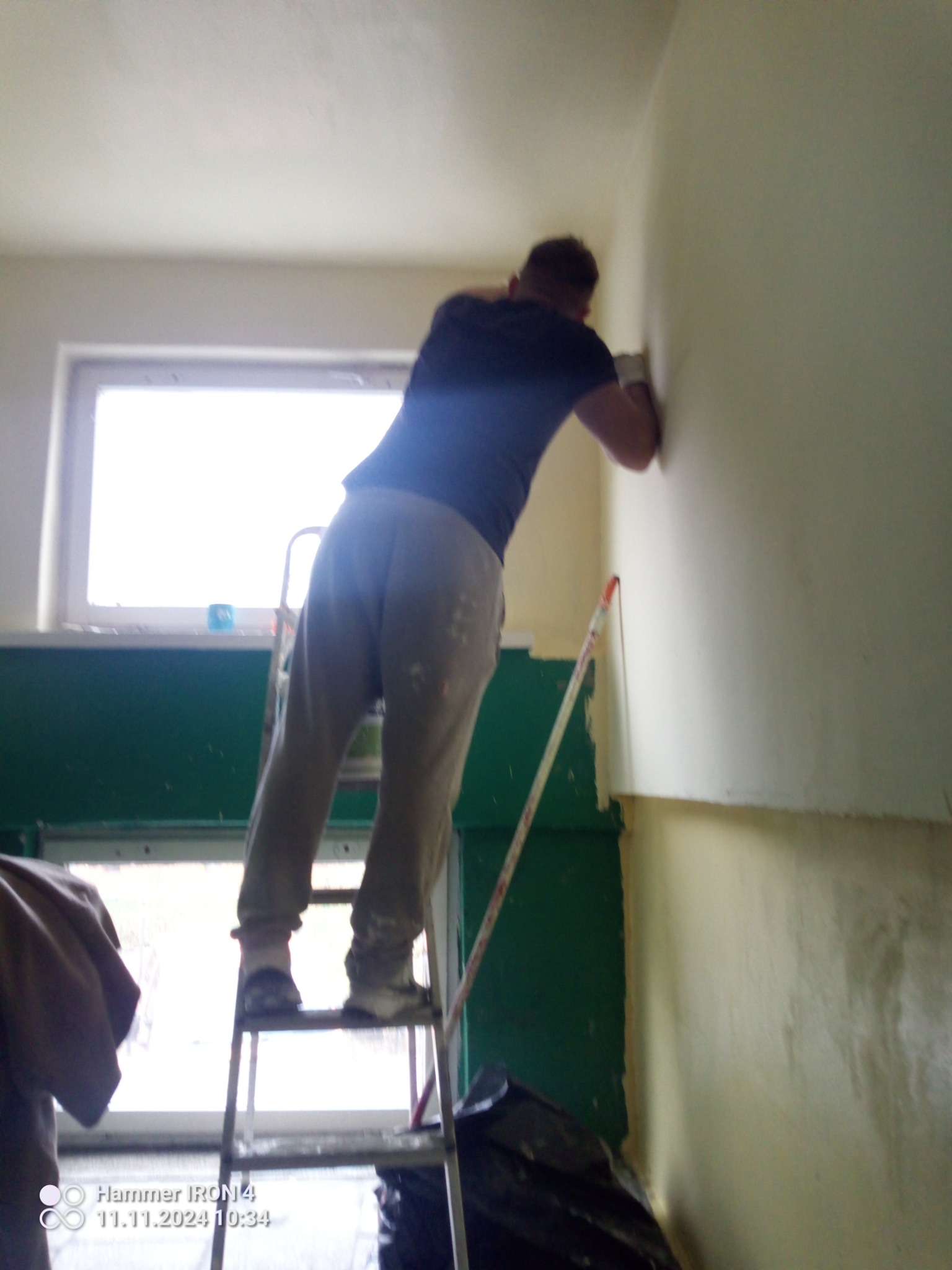 Mężczyzna na drabinie maluje ścianę wewnątrz budynku, widoczne niedomalowane fragmenty i świeża warstwa farby, w tle okno.