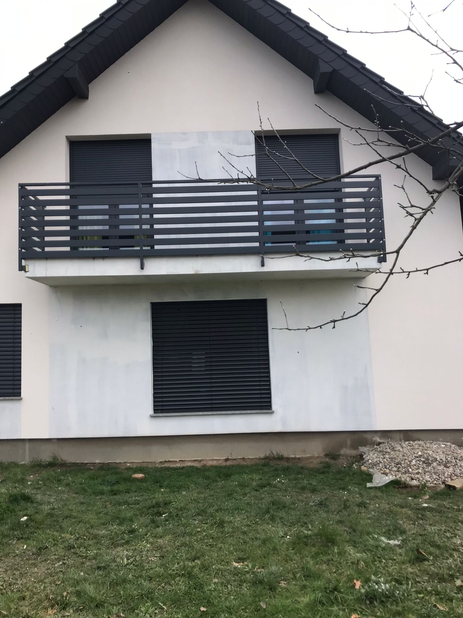 Biała elewacja domu z szarymi roletami i balkonem z metalową barierką, fragment elewacji przygotowany pod malowanie, widoczny stos kamieni na trawniku.