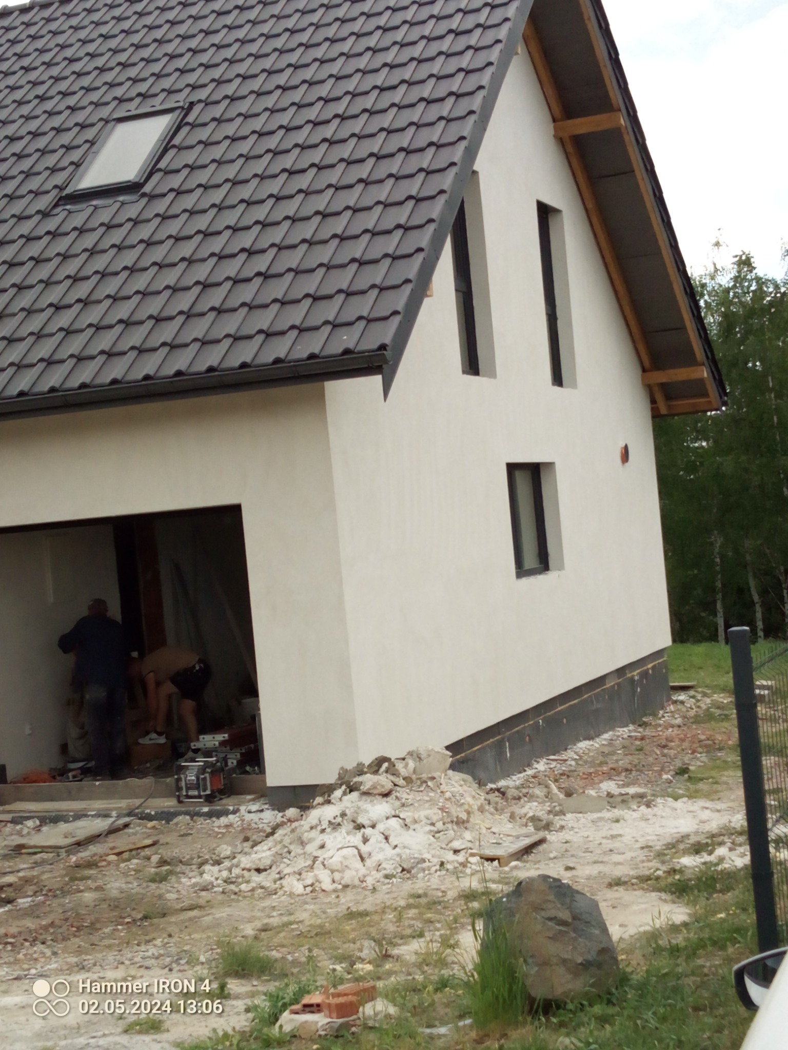 Nowo otynkowany dom jednorodzinny z ciemną dachówką, widoczni pracownicy w otwartym garażu, teren wokół domu w trakcie prac wykończeniowych.