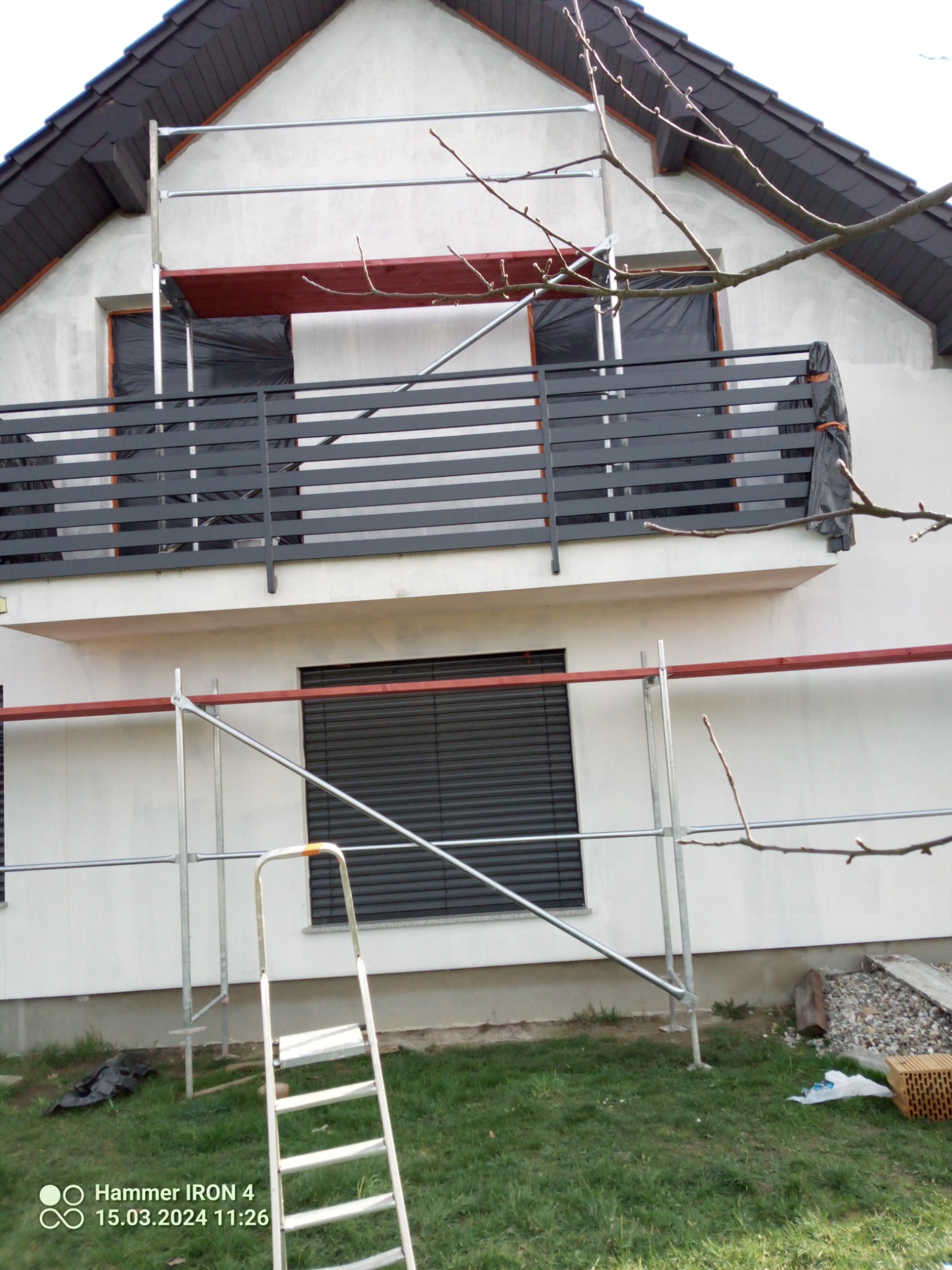 Dom jednorodzinny w trakcie prac elewacyjnych: widoczne rusztowanie, zabezpieczone okna folią, surowa elewacja, balkon z czarną balustradą, drabina na trawniku.