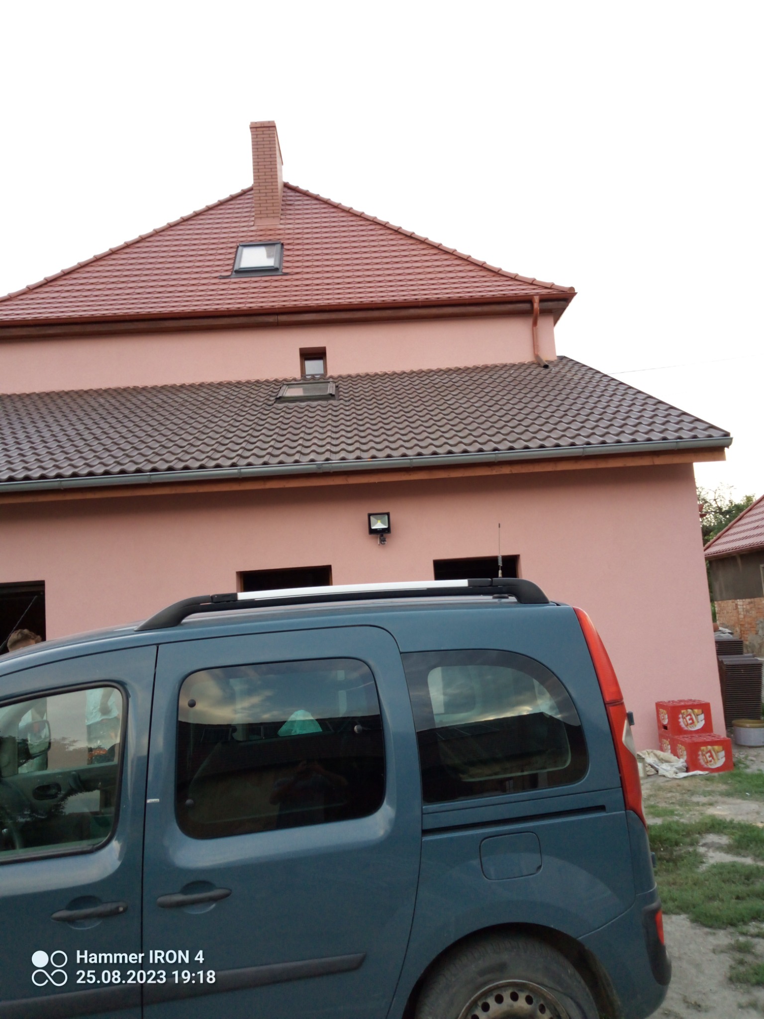 Dwukondygnacyjny budynek z różową elewacją i dwoma rodzajami dachówki, widoczny zza zaparkowanego, niebieskiego samochodu.