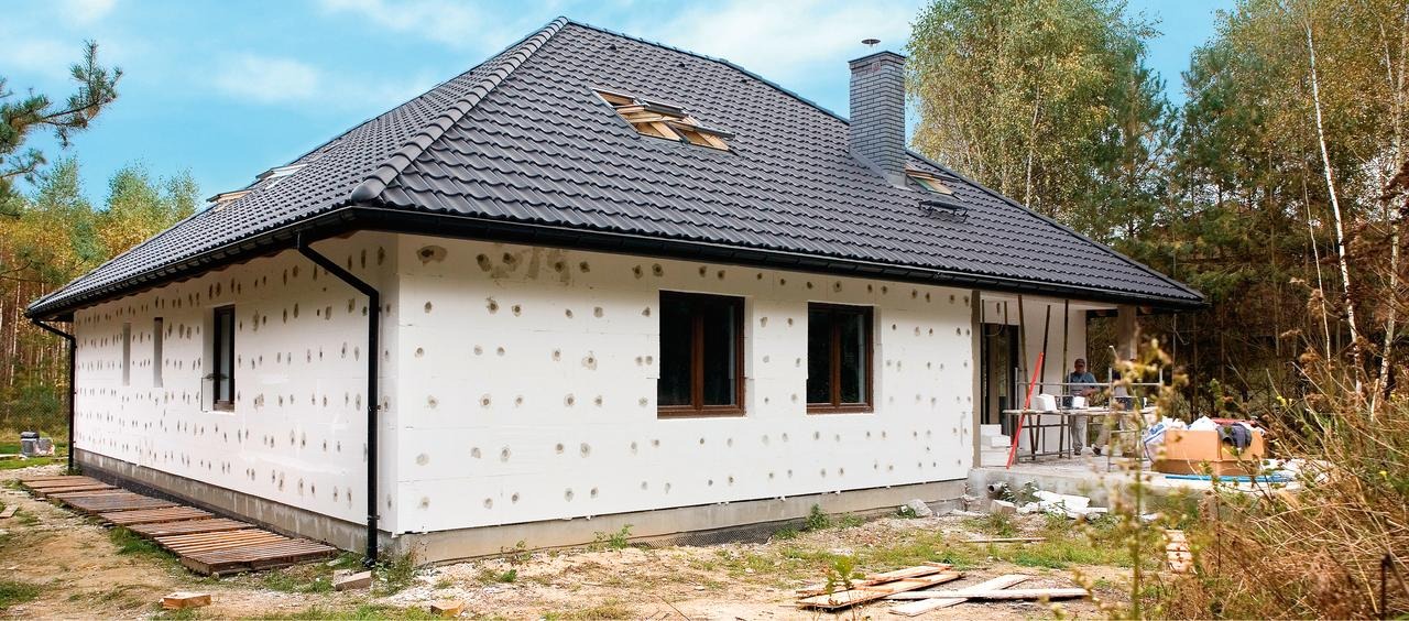 Dom jednorodzinny w trakcie ocieplania elewacji styropianem, widoczne otwory po kołkach mocujących, czarny dach z dachówki, otoczenie drzewa.