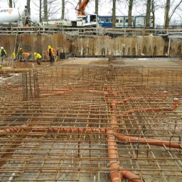Siatka zbrojeniowa z rurami kanalizacyjnymi w fundamencie, w tle pracownicy budowlani i maszyny na placu budowy.